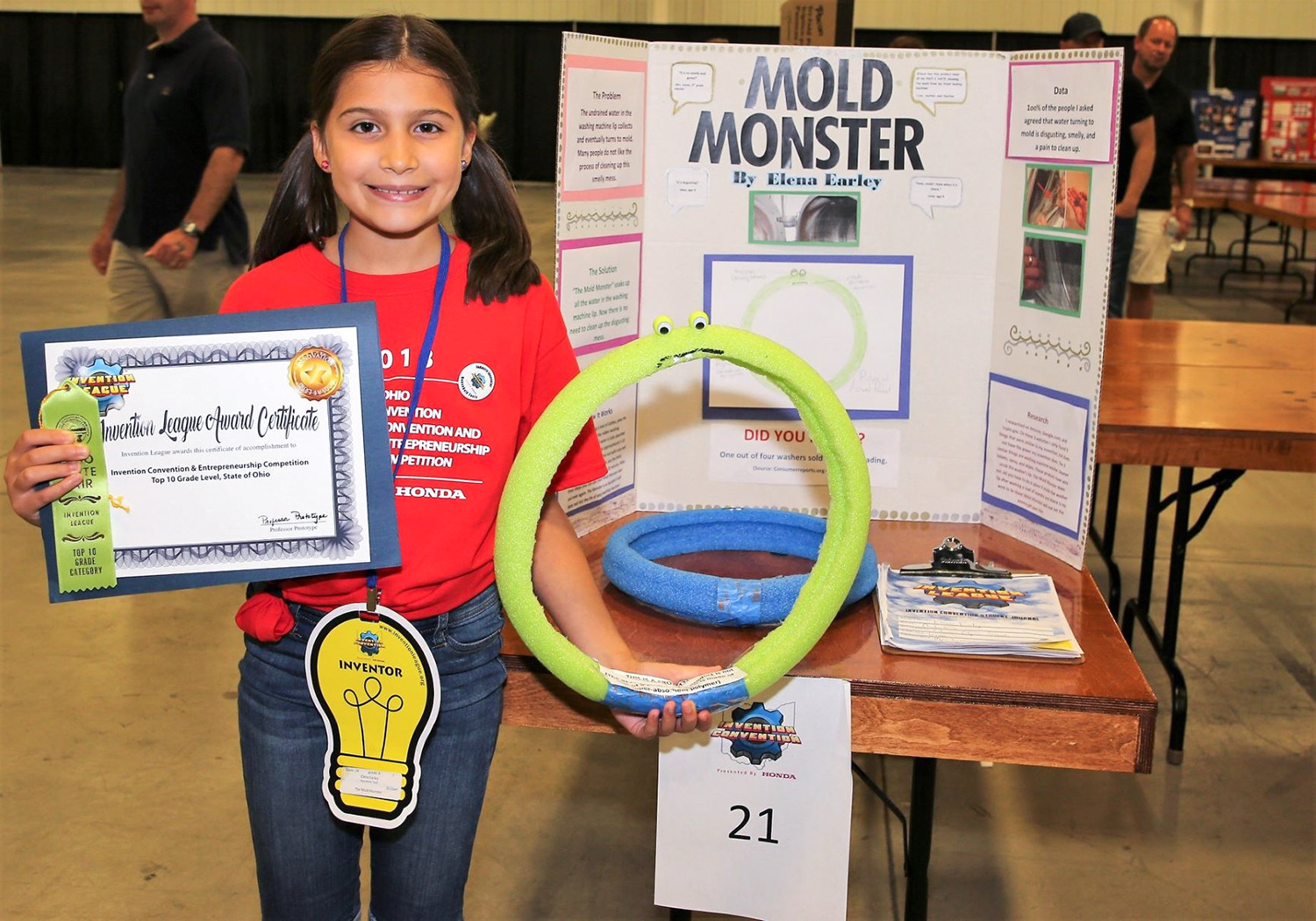 A young girl holds an award in one hand and a large green polymer ring in the other hand. She is standing in a gymnasium with a trifold informational poster board behind her with the words “mold monster” written in large black text. 