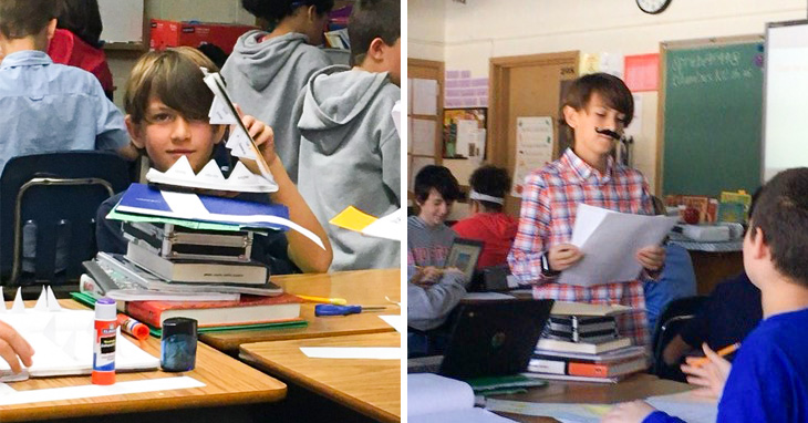 A young boy sits at a school desk with books piled on top as he peers through a book with shark-like teeth. A boy wearing a plaid shirt and fake mustache standing up in a classroom while delivering an oral presentation.