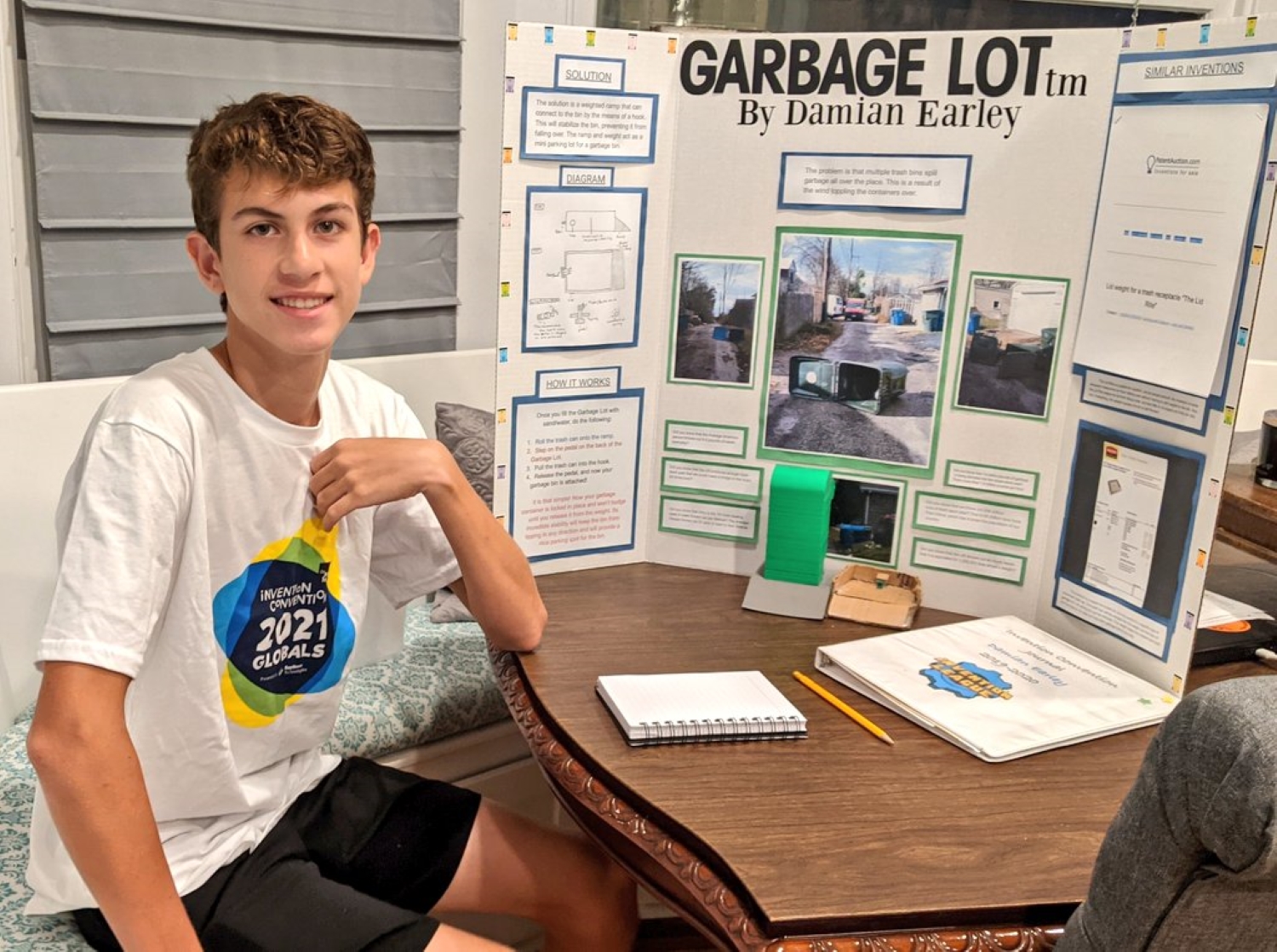 A male high school student wearing a white t-shirt sits at a table with a trifold poster board in the background detailing his garbage can platform invention.