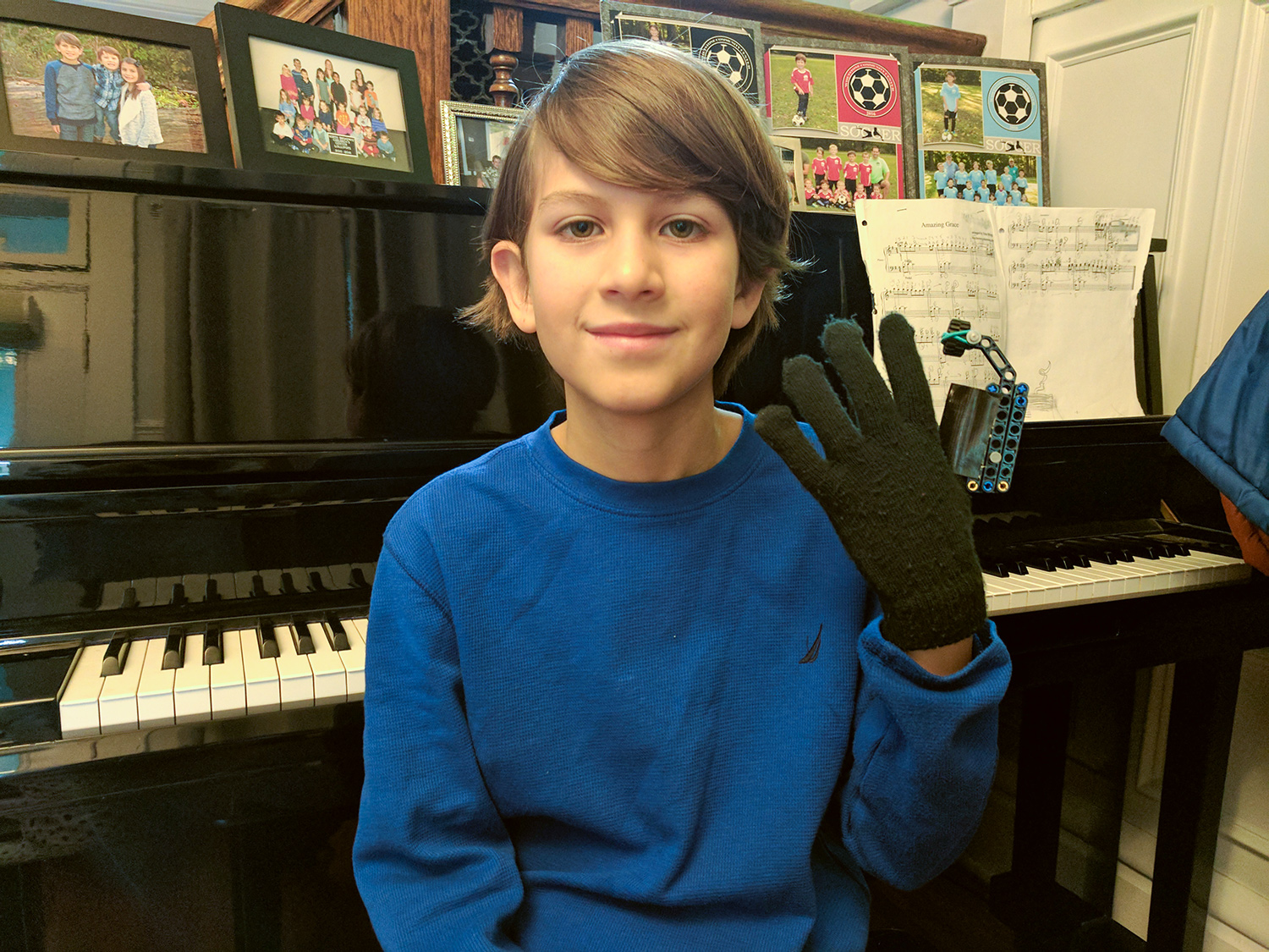 A fifth-grade boy wearing a blue sweater sitting on a piano bench wearing a glove that has a mechanical extension to increase the finger-reach span for pianists.
