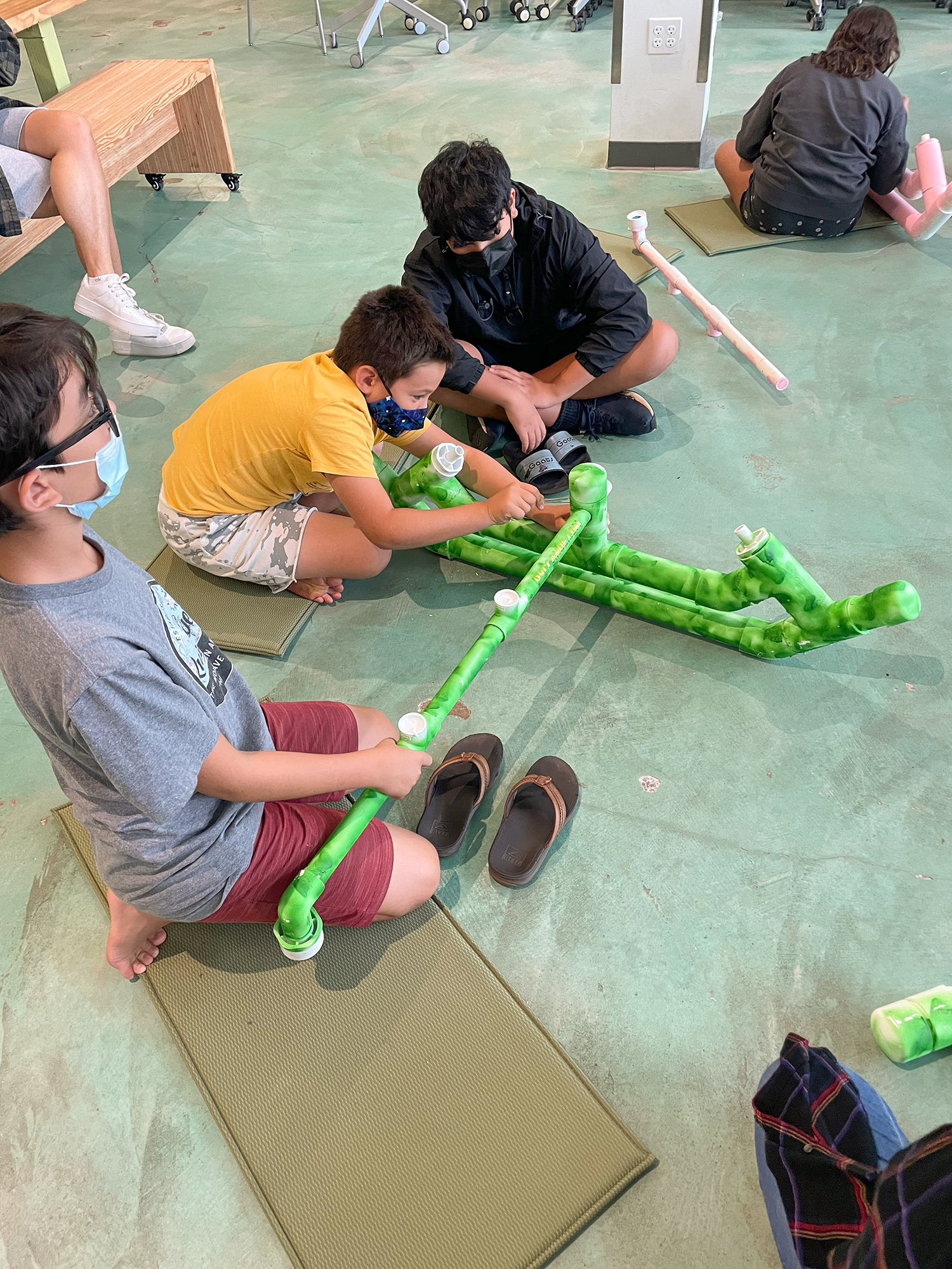 Three school-age children are sitting on the floor working together to build a canoe prototype our of PVC pipe.