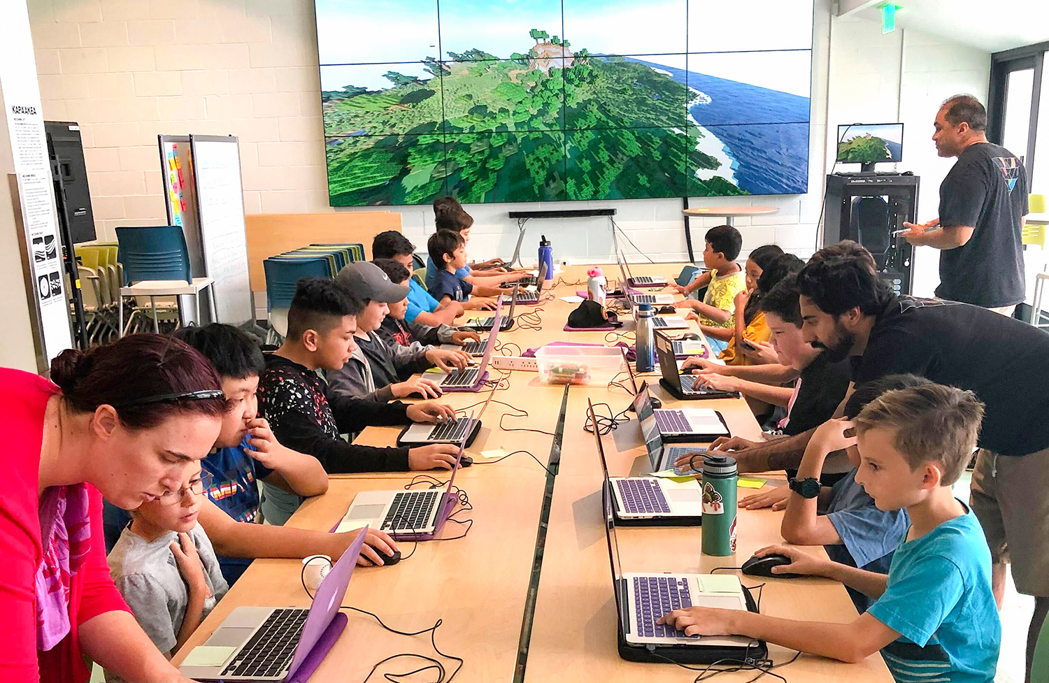 School-age children are sitting at a rectangular table with computers in front of them as they learn to code using a popular online game called Minecraft. 