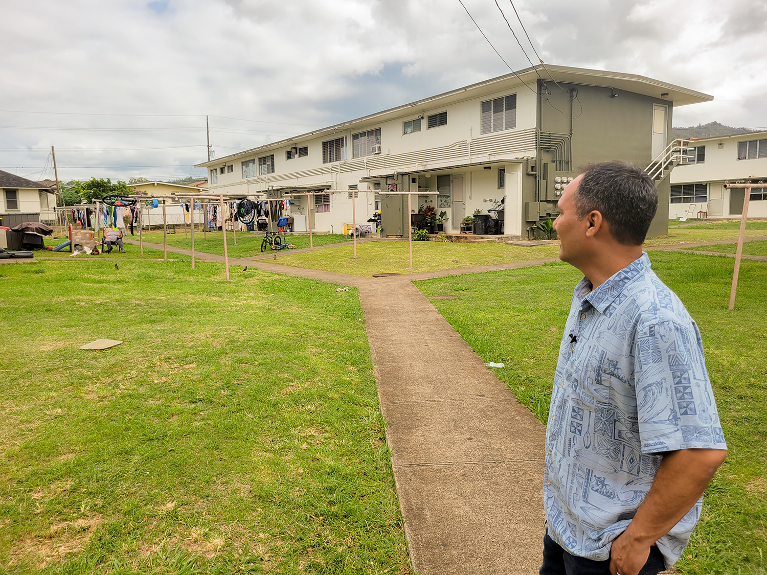 A man in a blue shirt stands looking away from the camera at a two-story multi-family housing unit. 