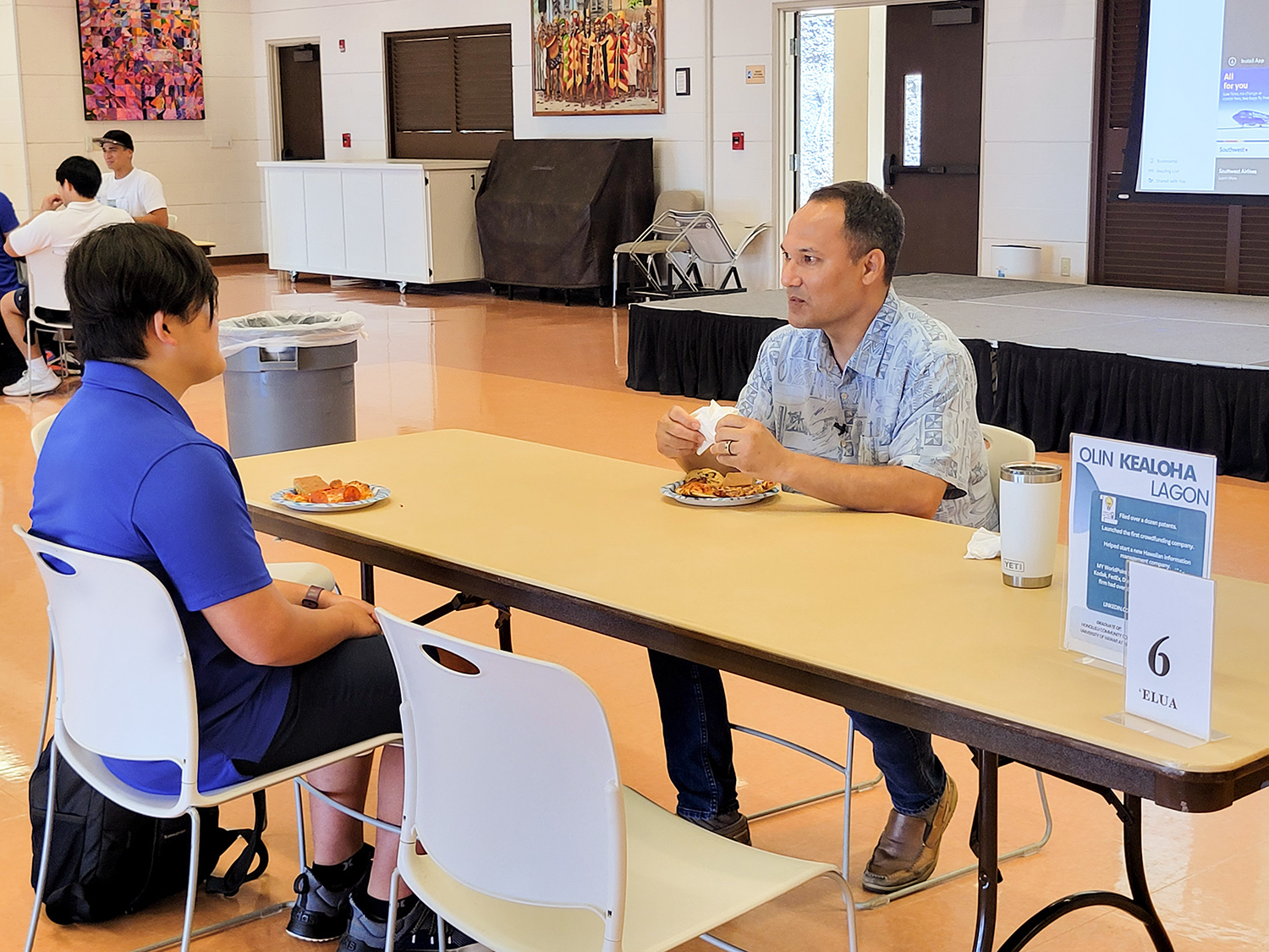 Man in a blue shirt is seated at a rectangular table and is talking to a male high school student sitting across from him.
