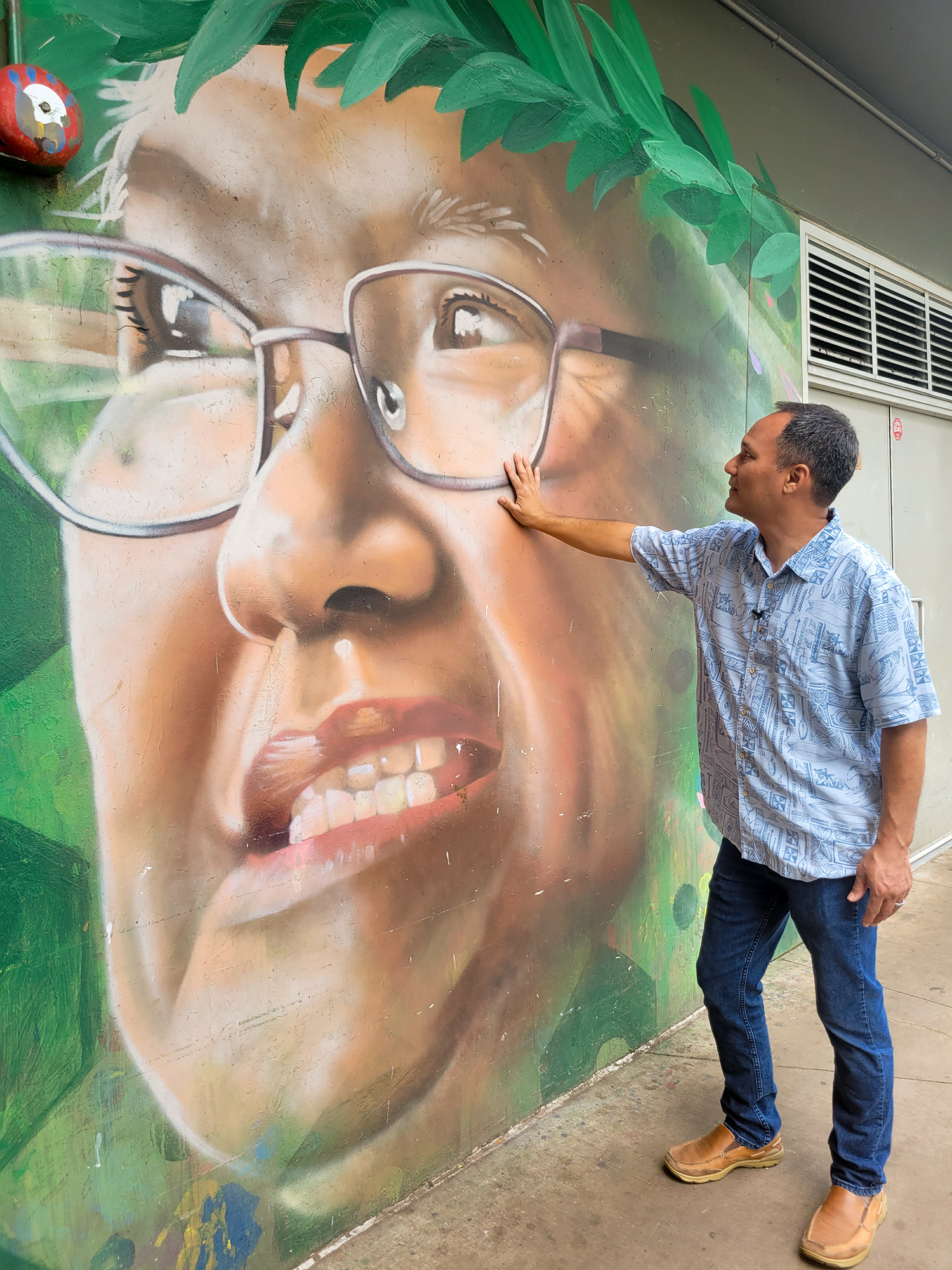 Man in a blue shirt and jeans is standing near a wall with a mural painted on it. The mural is the face of the man’s mother. He has his hand placed on the mother’s cheek. 