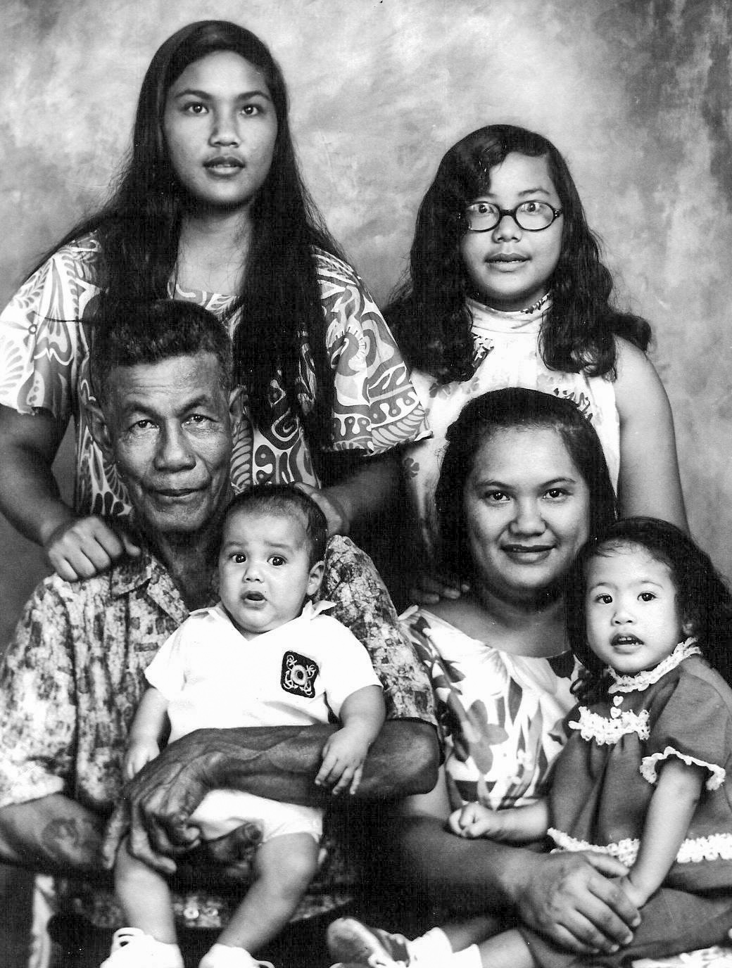 A black and white photograph showing a family seated and standing closely together. Photographed are two adults, seated, a young boy on the man’s lap, a young girl sitting on the woman’s lap and two teenage girls standing behind the adults. 