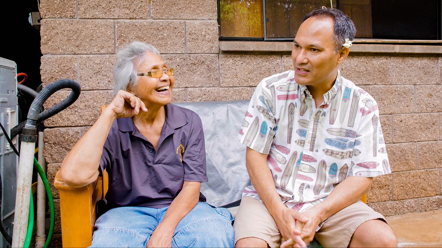 A man in khaki shorts and surfboard button-up shirt sits next to an elderly woman as they discuss energy costs in Hawaii.