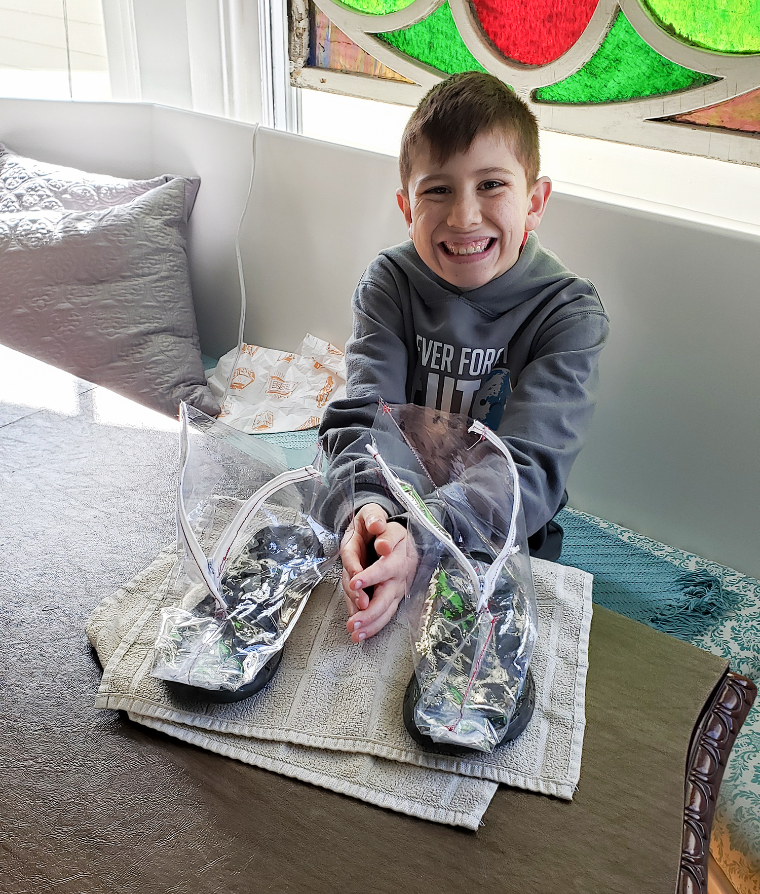 Young boy sitting down at a table with his invention, clear vinyl shoe protectors, displayed.