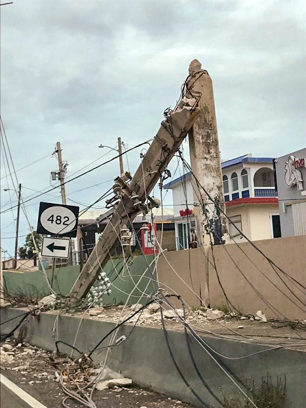 A collapsed concrete utility pole is seen bordering a highway in Puerto Rico, broken in half with the lower half still standing. Power lines lie on the ground.