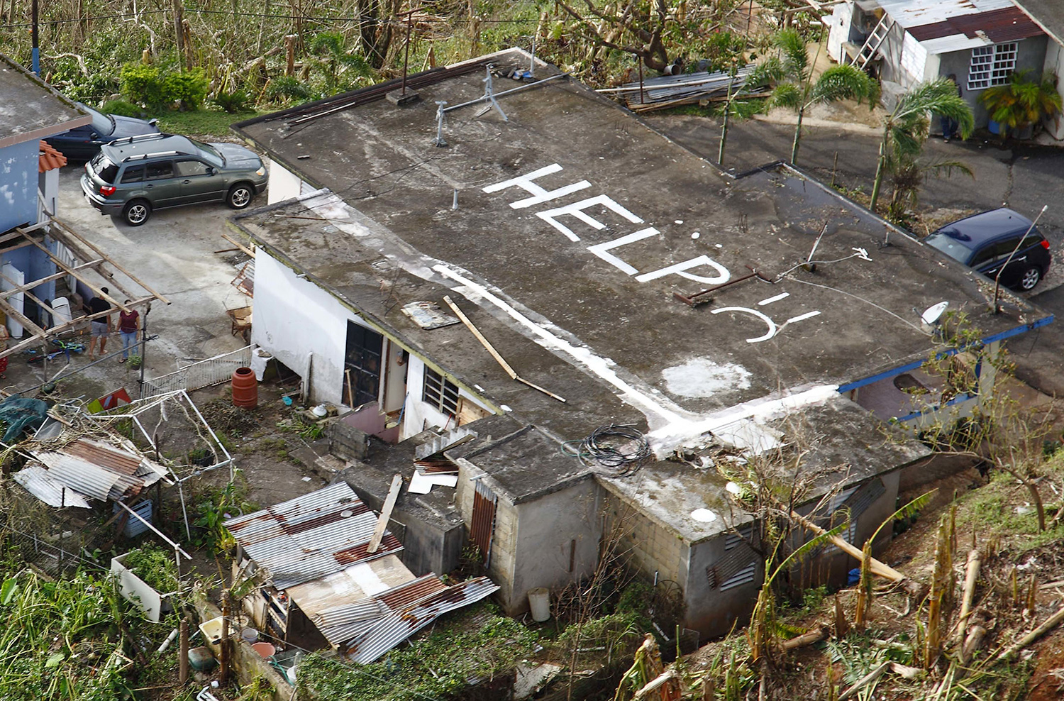 A house in Puerto Rico is seen from the air. Someone has written “Help” in white on the roof for rescue airplanes and helicopters to see. There is debris and fallen vegetation around the house.