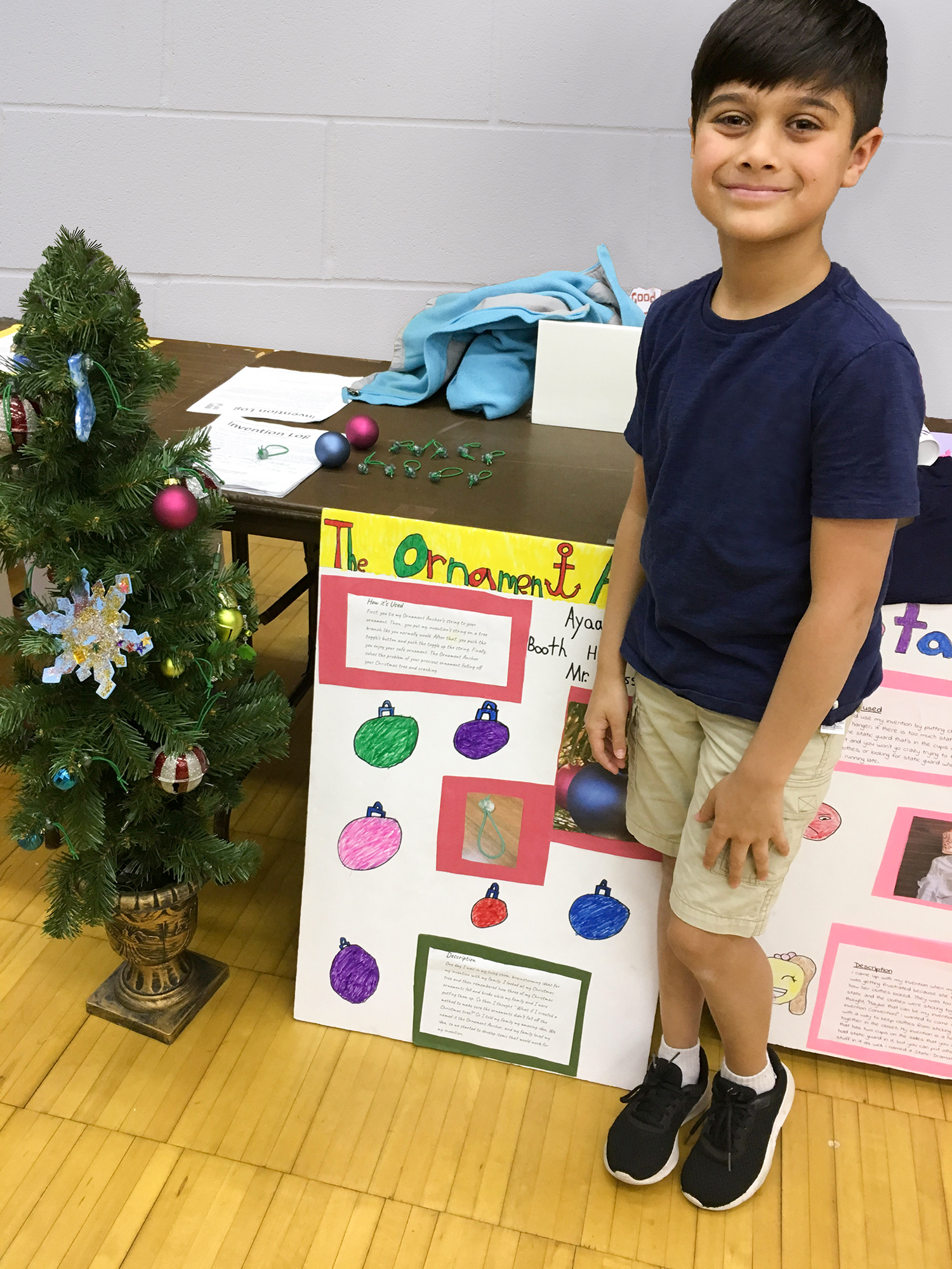 A boy stands next to his project about Ornament Anchor. Near him is a small Christmas tree with ornaments, a poster display and a table with ornaments.