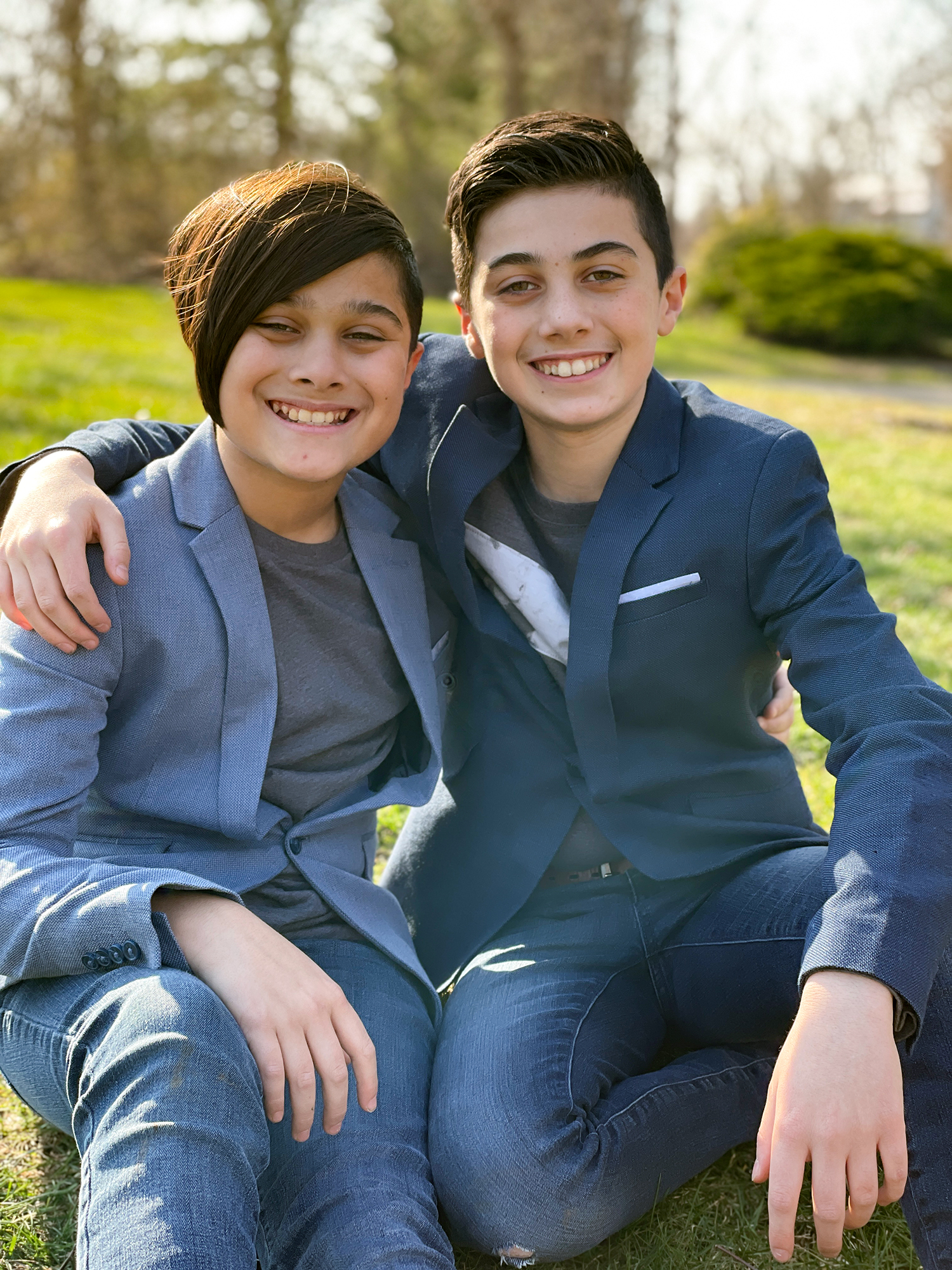Two teenage brothers dressed in business suits sit next to each other. The older brother has his arm around the shoulder of the younger