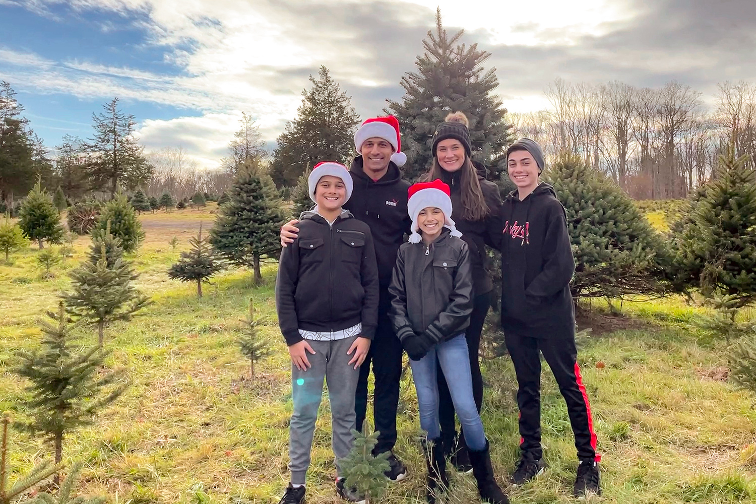 A family of a mom, dad, girl and two boys stand in a field with a background of Christmas trees.