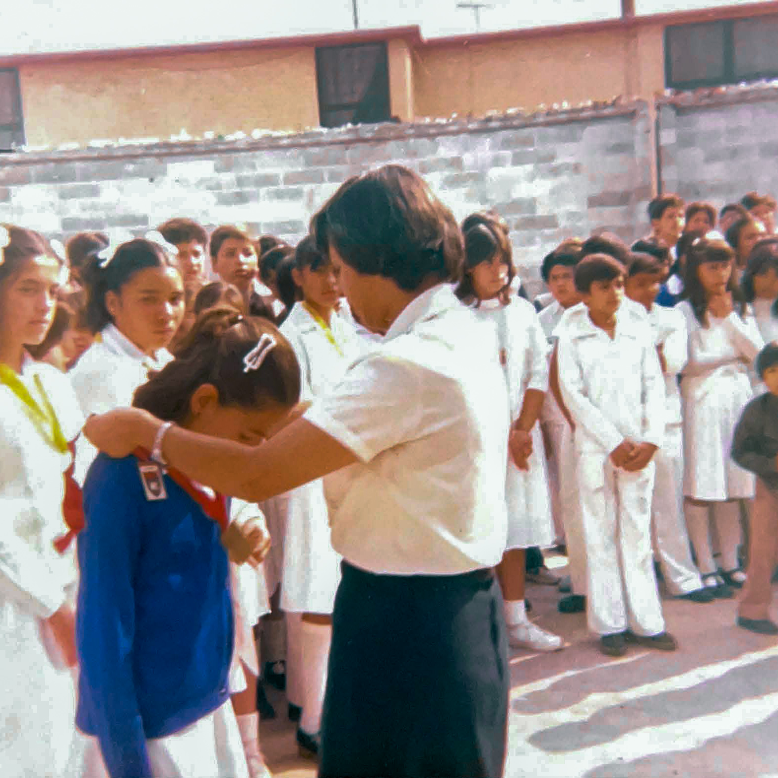 Una niña de escuela primaria con un cárdigan azul se inclina para aceptar una medalla alrededor de su cuello que le entrega una mujer con una blusa blanca y una falda oscura.