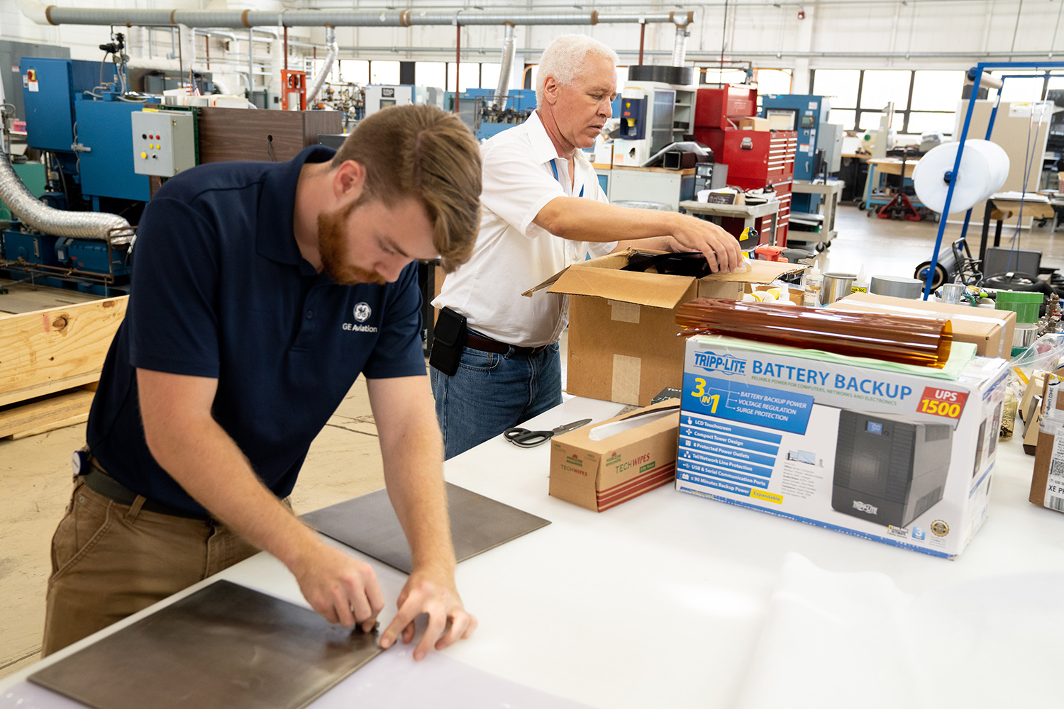 A man with light brown hair and beard wearing a navy blue polo and brown pants works with a sheet of metal while in the background Robert Bryant in a white button up shirt and jeans unboxes supplies