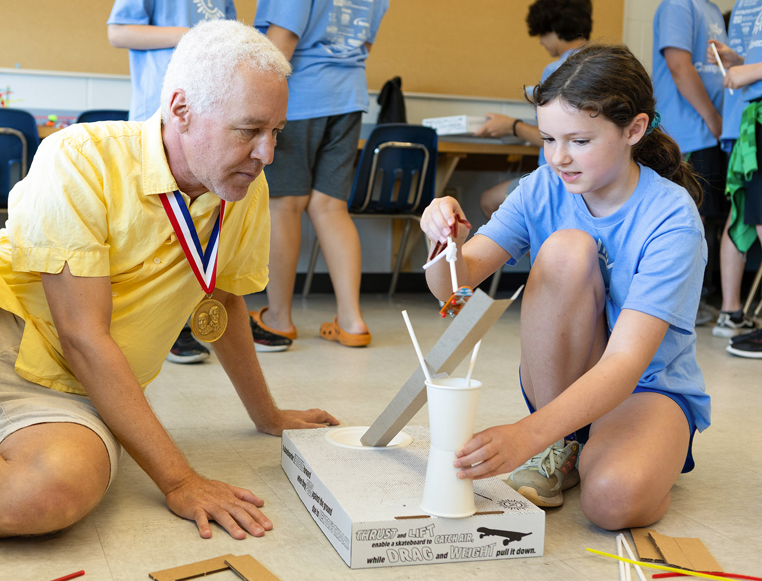 Robert Bryant in a yellow polo shirt and khaki shorts with a National Inventors Hall of Fame medal around his next observes a young girl with brown hair, blue shirt, and blue shorts explain her project in a classroom