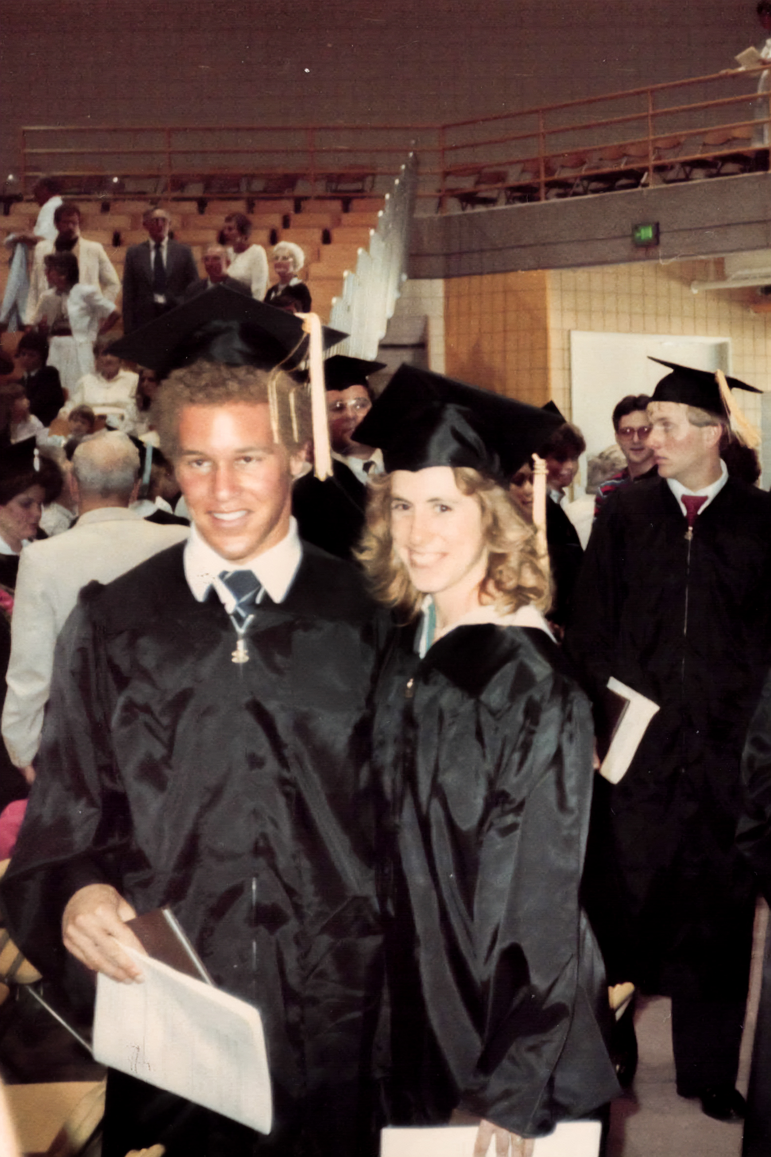 Robert Bryant and his future wife Joy pose in black graduation caps and gowns in crowded gymnasium with other graduates and attendees