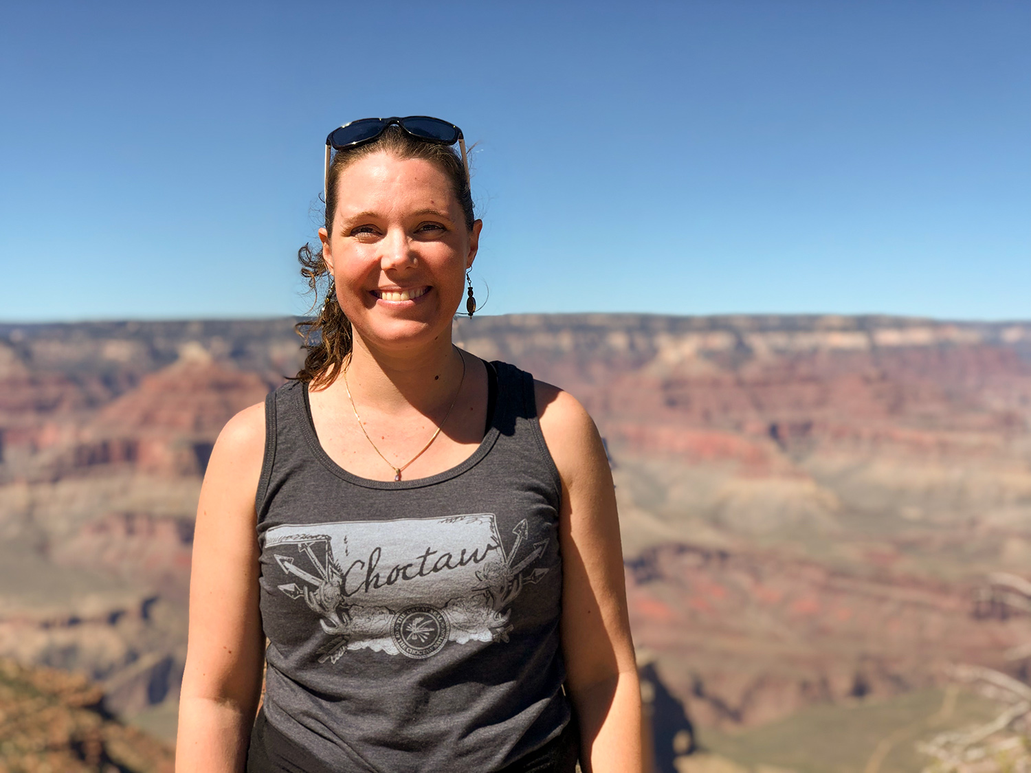 Tara Astigarraga stands in front of the red and green cliffs of the Grand Canyon, smiling and facing the camera. She wears sunglasses on the top of her head and a gray and white tank top with an outline of the state of Oklahoma and the words “Choctaw” written in cursive.