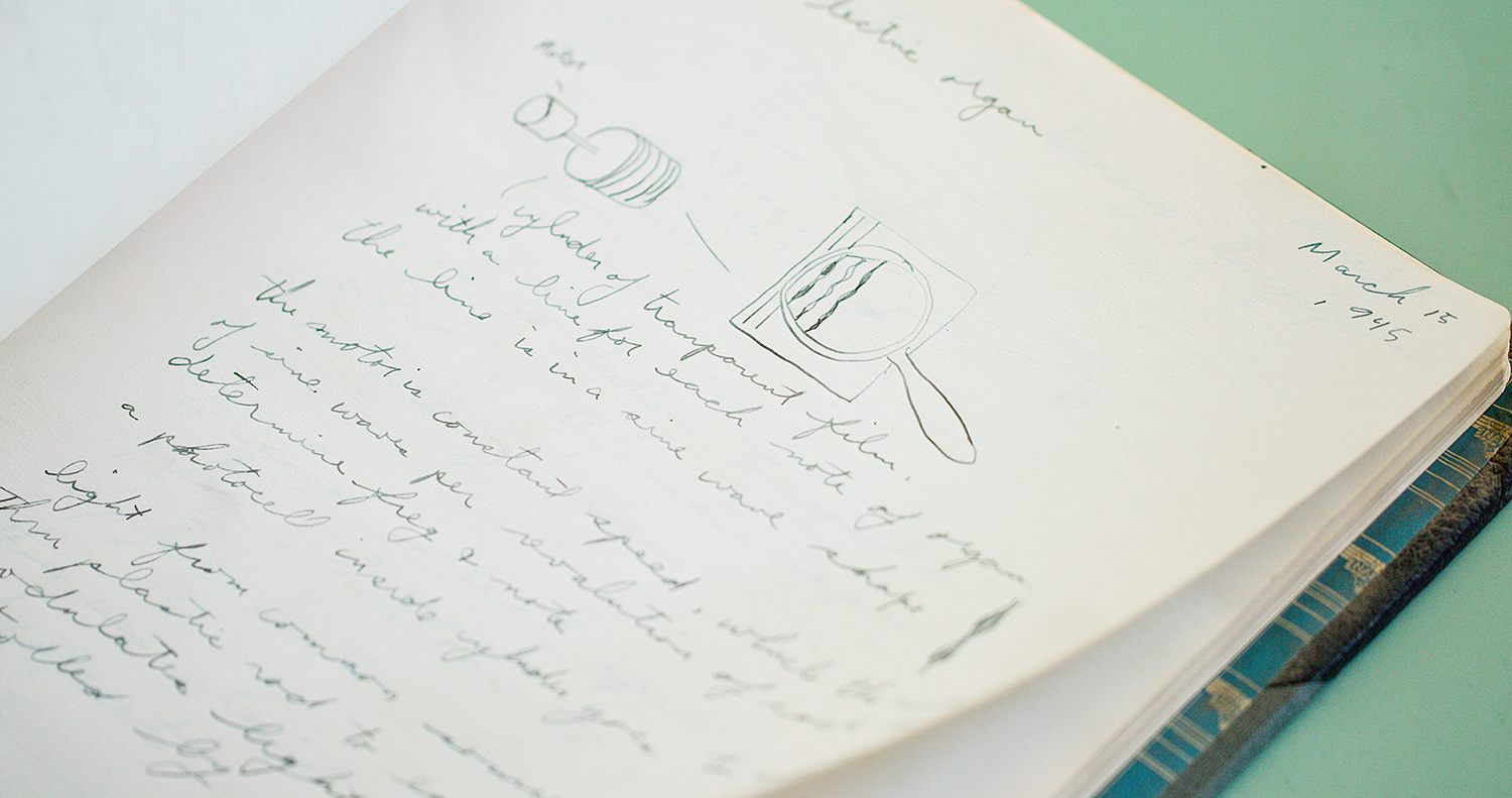 An open notebook containing design sketches and cursive handwriting written in pencil is shown on white paper.