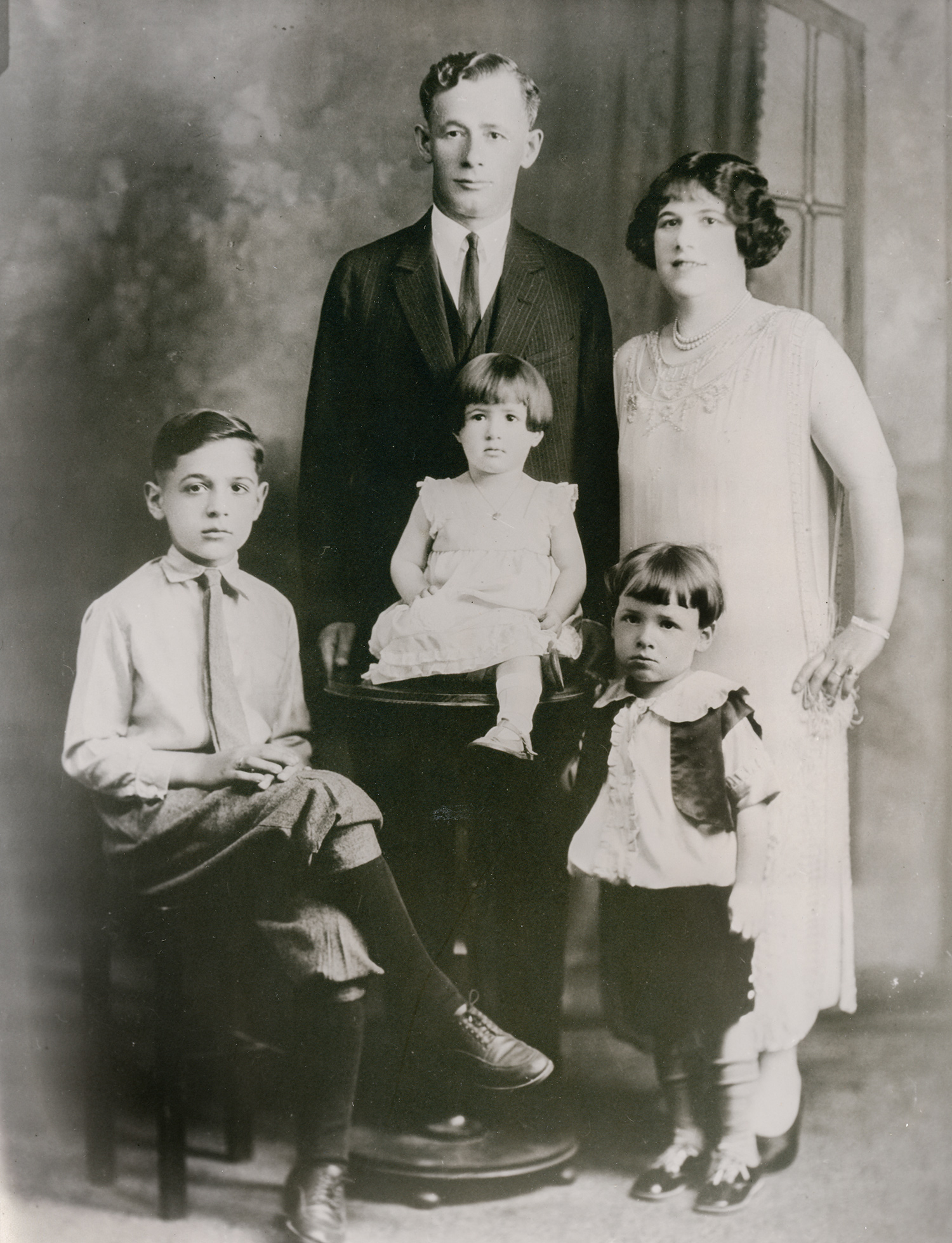 A family of two adults and three children pose in early 20th century attire. A man wearing a dark suit stands next to a woman wearing a light-colored long dress in the background while their children, a school-age male, a toddler female, and a young male child pose in the foreground. 