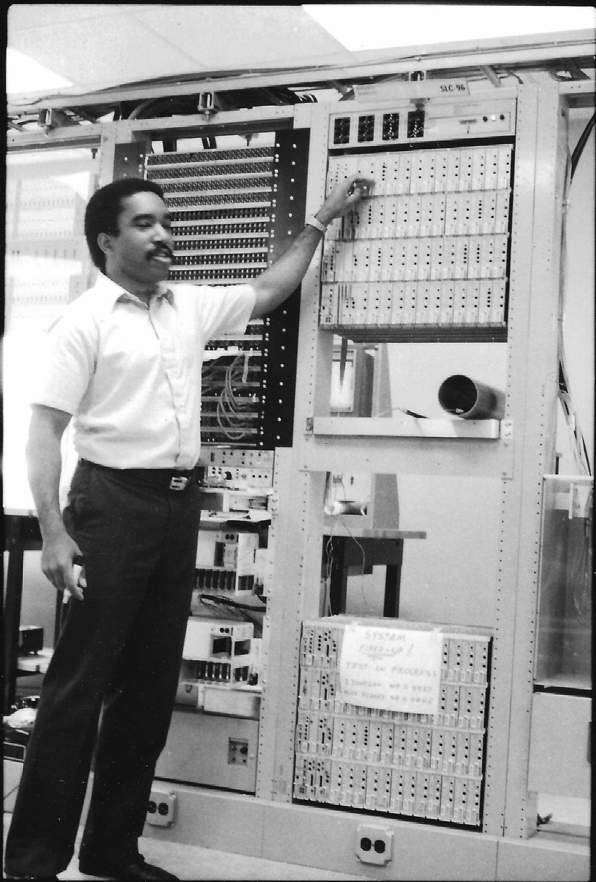 Lanny Smoot standing next to and point at a fiber optic system at Bell Labs. A sign on the system says “System fired up! Test in progress”