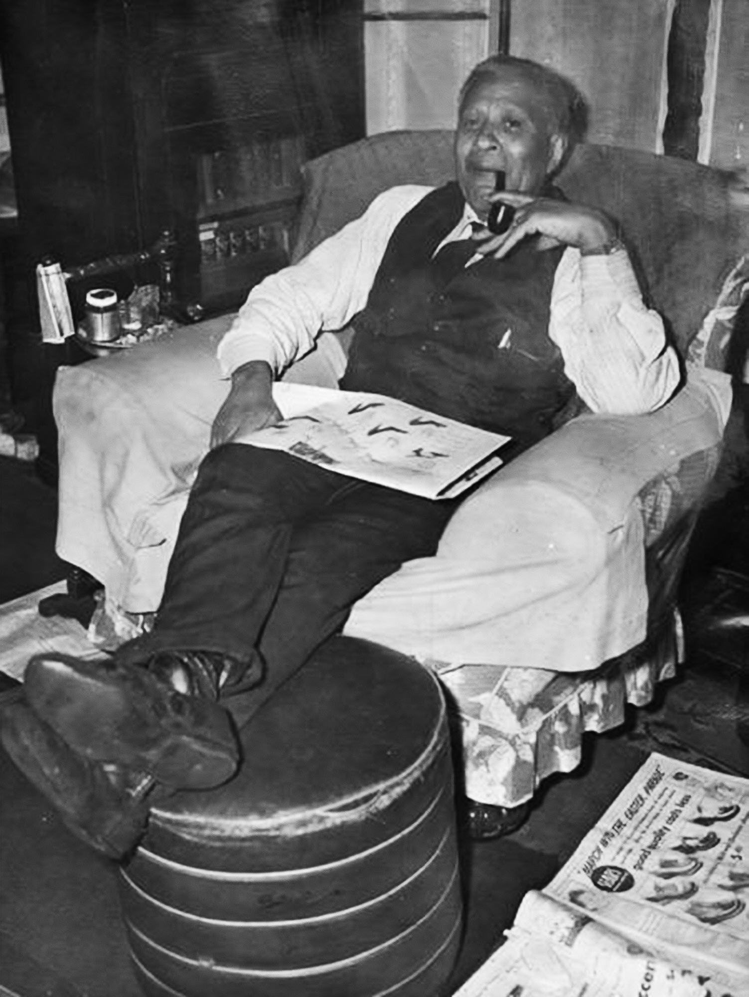 Garrett Morgan, in a vest and tie with a newspaper on his lap, relaxes in an armchair while smoking a pipe