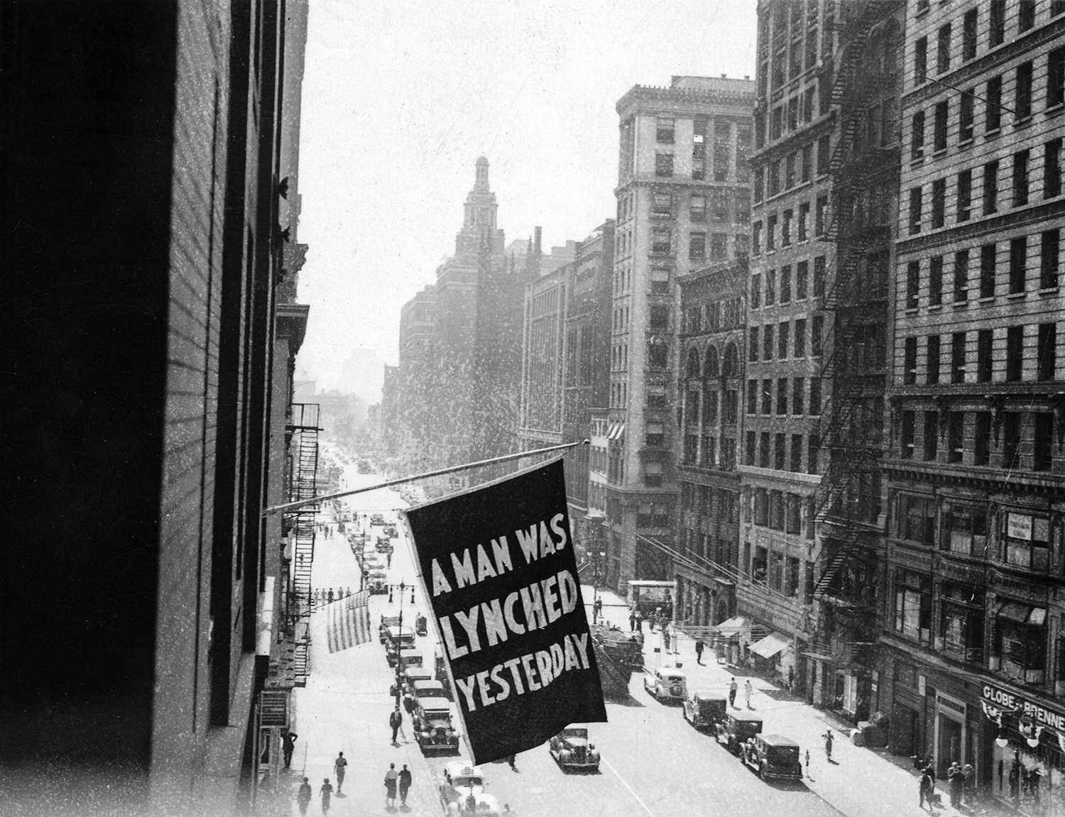 A window flag reading “A man was lynched yesterday” in bold letters in the foreground with a busy New York City street with buildings on either side in the background