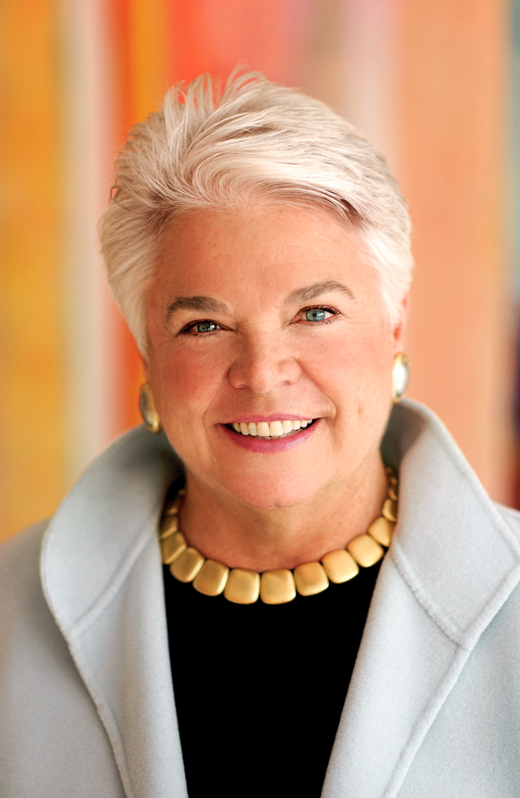 A recent portrait of entrepreneur and author Pleasant T. Rowland. Rowland smiles engagingly at the camera, while gold jewelry and a white jacket with popped color and dark blouse accentuates her bright white hair.