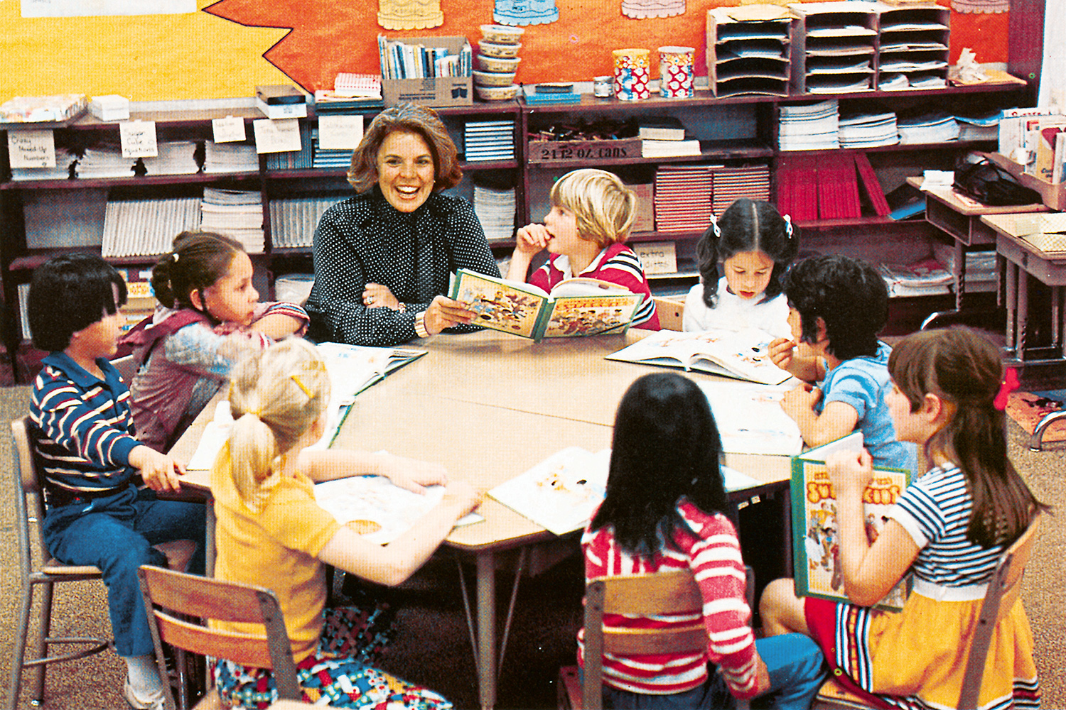 Pleasant Rowland smiles delightedly as a diverse group of young children gathered at a classroom table read from the textbook she created.