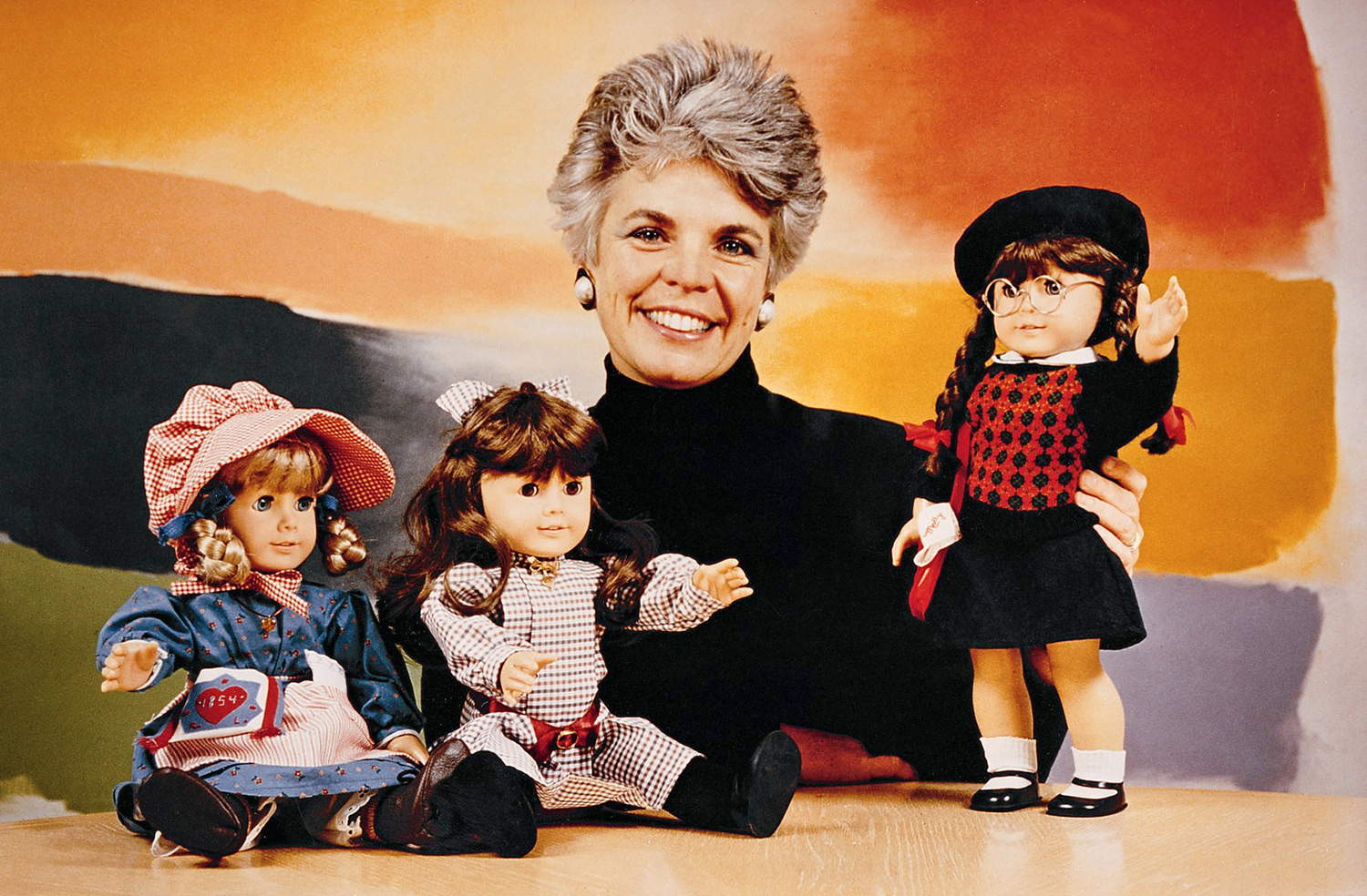 American Girl doll creator Pleasant T. Rowland smiles alongside the three first American Girl Dolls launched in 1986. The dolls wear historically accurate outfits. At left is Kirsten, in a pioneer bonne and apron, next to her is the Samantha doll in a plaid turn of the century dress, then Rowland herself smiling brightly at the camera with arms crossed and wearing a black turtleneck, and on the far right is Molly dressed in 1940s bobby socks, argyle sweater and skirt.