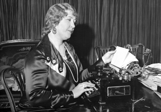 A middle-aged woman in a silk dress sitting in a chair while examining a typewriter