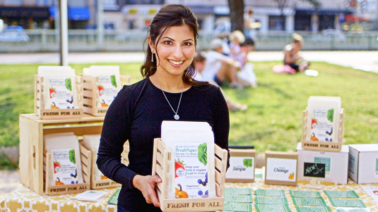 Kavita Shukla holding sheets of FreshPaper in a carton that says “fresh for all” at a farmers market in front of a table advertising FreshPaper 