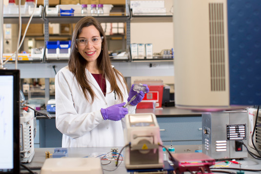 Daniela Blanco, a brown-haired young Latino woman, wears a white lab coat, safety glasses, and plastic gloves as she stands amid chemistry equipment in a well stocked lab. She pauses her experiment momentarily to smile at the camera.