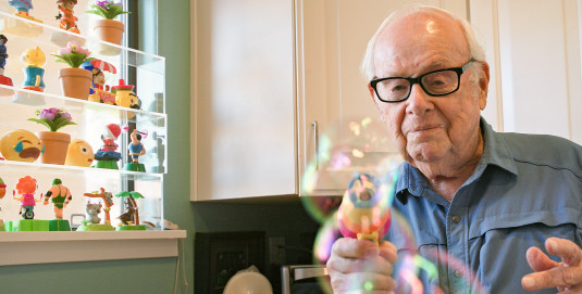 An elderly man wearing a blue collared shirt and black eyeglasses blows bubbles towards the camera using a battery-powered bubble gun.