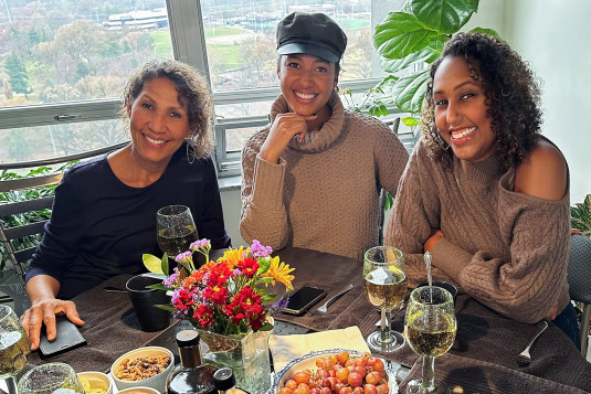 Three women with dark tan skin, brown hair, and long neutral-colored long sleeve tops smiling at a dinner table set with food, wine, and flowers.