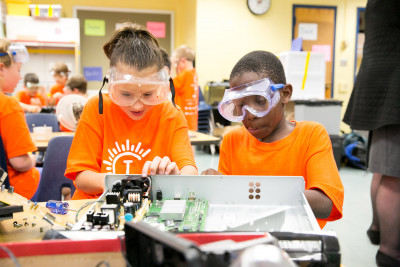 Two Camp Invention students wearing safety googles work on disassembling electronics.