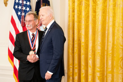 Neil Gilbert Siegel wearing a dark suit and glasses shakes President Biden’s hand, after receiving the National Medal of Technology and Innovation.