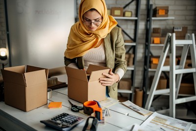 A person in a Hijab is smiling and packaging cardboard boxes in a mail room.