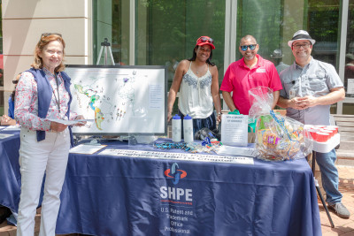 Two women (one in white jeans, one in a tank top and hat) and two men (one in sunglasses, one in a hat) smile while standing outdoors around a table with a blue SHPE USPTO tablecloth, with giveaways on the table.