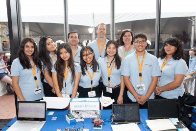 a diverse group of 10 adults and young inventors smiling and standing behind a table with three laptops and the invention