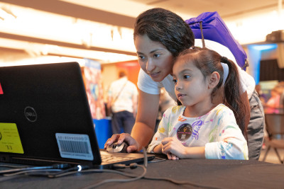 A young girl with long dark brown pigtails and dark eyes smiles while looking at a laptop screen on a table at USPTO headquarters. Her caretaker, a woman with dark hair and dark eyes, leans over her shoulder, also looks at the screen, and clicks a computer mouse.  