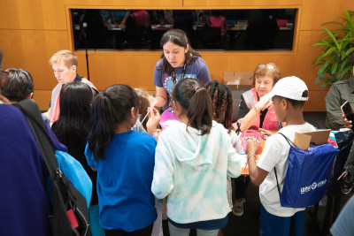 At USPTO headquarters, a woman with dark hair wearing a lanyard is surrounded by seven diverse children who are gathering pipe cleaners and beads from her craft table.