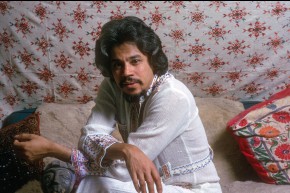 Johnny Pacheco sits on cushions and faces the camera. He wears an embroidered linen shirt.