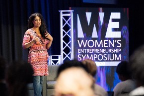female standing on a stage next to a sign for 'Women's entrepreneurship symposium'. blurry heads in the foreground of the event.