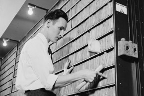 Black and white photo of a man in a dress shirt and tie searching through hand-written records in a row of shelves