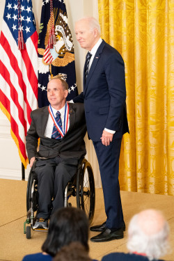 Rory Cooper in a dark suit and wheelchair poses with President Biden after receiving the National Medal of Technology and Innovation.