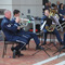 Photo of five man band sitting in chairs playing the trumpet, trombone and tuba.