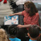 Photo of a woman sitting and reading in front children with Cat in the Hat standing behind her.